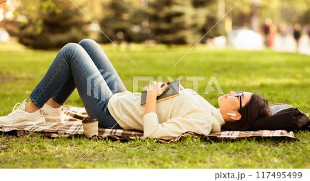 Asian young woman wearing casual clothes is lying on a blanket, holding a book, and listening to music with wireless earbuds. She is surrounded by lush green grass and trees on a sunny day 117495499
