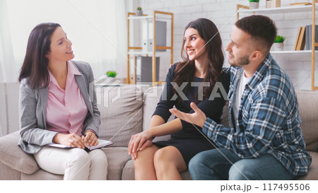 A young couple is sitting on a couch in an office setting, talking with their therapist. The therapist is a woman, and she is listening intently to the couple A young couple is sitting on a couch in an office setting, talking with their therapist. The therapist is a woman, and she is listening intently to the couple 117495506