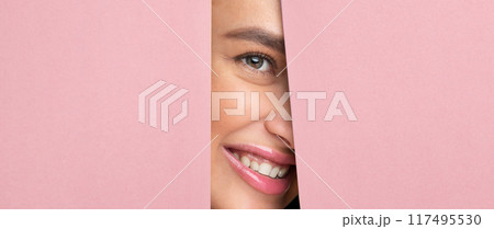Cheerful woman with her hair in a bun and wearing a white shirt is peeking out of a hole in a vibrant pink wall. Cheerful woman with her hair in a bun and wearing a white shirt is peeking out of a hole in a vibrant pink wall. 117495530
