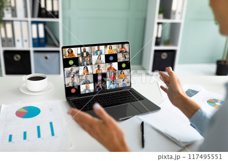 A person sits at a desk in an office setting and leads a video conference on their laptop. The laptop screen shows a grid of many video feeds, showcasing a diverse group of people A person sits at a desk in an office setting and leads a video conference on their laptop. The laptop screen shows a grid of many video feeds, showcasing a diverse group of people 117495551