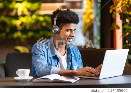 A young black guy is seated in a coffee shop, focused on using his laptop. He is typing on the keyboard, surrounded by tables and chairs. The background shows other customers and baristas. 117495603