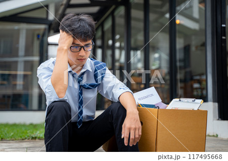 Sad young businessman sitting at a curve side road with a box full of his office stuff beside him Sad young businessman sitting at a curve side road with a box full of his office stuff beside him 117495668