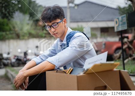 Sad young businessman sitting at a curve side road with a box full of his office stuff beside him 117495670