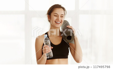 A woman standing while holding a water bottle in one hand and a yoga mat in the other hand. Healthy lifestyle concept 117495798