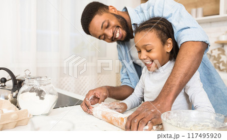 A cheerful african american father teaches his daughter how to handle dough in their kitchen, emphasizing the joys of fatherhood and learning A cheerful african american father teaches his daughter how to handle dough in their kitchen, emphasizing the joys of fatherhood and learning 117495920