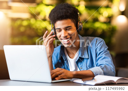 African American young guy is seated, wearing headphones and focused on using a laptop. He appears engrossed in his work, typing and clicking with concentration. African American young guy is seated, wearing headphones and focused on using a laptop. He appears engrossed in his work, typing and clicking with concentration. 117495992