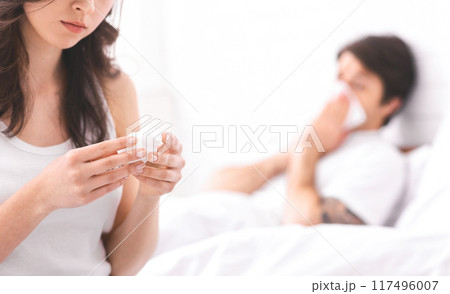 This image shows a woman holding napkin, while her partner lies in bed, visibly sick and using a tissue to blow their nose. Loving relationship and a caring attitude towards a sick partner. This image shows a woman holding napkin, while her partner lies in bed, visibly sick and using a tissue to blow their nose. Loving relationship and a caring attitude towards a sick partner. 117496007