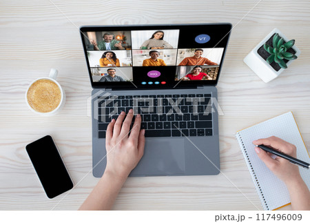 A woman is participating in a video conference meeting on her laptop. She is typing on the keyboard with one hand and writing notes with the other. A woman is participating in a video conference meeting on her laptop. She is typing on the keyboard with one hand and writing notes with the other. 117496009