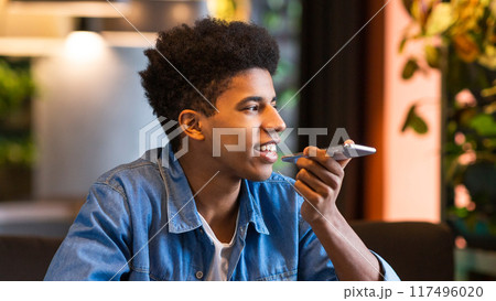 A young black guy is sitting in a modern cafe, holding his smartphone and using a voice assistant to perform tasks or send messages. A young black guy is sitting in a modern cafe, holding his smartphone and using a voice assistant to perform tasks or send messages. 117496020