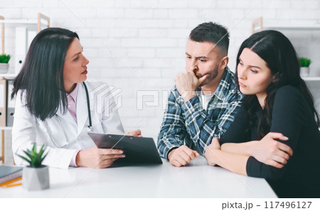 A doctor sits at a table with a young couple. The doctor is holding a clipboard and appears to be talking with the couple. The couple looks concerned and listens intently to the doctors advice. 117496127