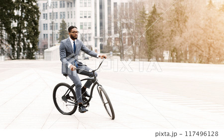 Black businessman is biking in an urban environment. The mood is energetic and productive, capturing the essence of a modern professional on the go. 117496128