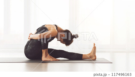 A woman is performing a yoga pose on a mat in a bright studio. She is stretching her legs and back, demonstrating flexibility and focus. A woman is performing a yoga pose on a mat in a bright studio. She is stretching her legs and back, demonstrating flexibility and focus. 117496163