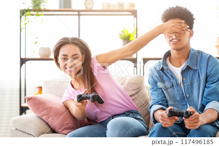 African American girl is covering her boyfriends eyes with her hand while he plays video games on the couch. They are both smiling and having fun. They are likely in a living room or home setting. African American girl is covering her boyfriends eyes with her hand while he plays video games on the couch. They are both smiling and having fun. They are likely in a living room or home setting. 117496185