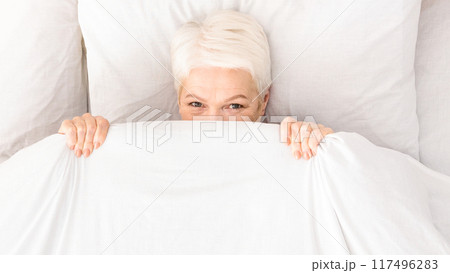 Playful senior woman in bed hiding under blanket, top view, panorama with free space Playful senior woman in bed hiding under blanket, top view, panorama with free space 117496283