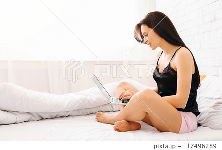 A woman is seated on a bed, engaging with a laptop computer. She appears focused and concentrated on the screen as she types or reads. The room is softly lit, creating a cozy atmosphere. 117496289