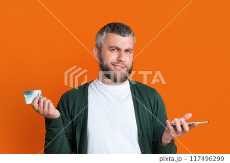 A bearded man stands against an orange background, holding a credit card in one hand and a smartphone in the other. He looks puzzled. 117496290