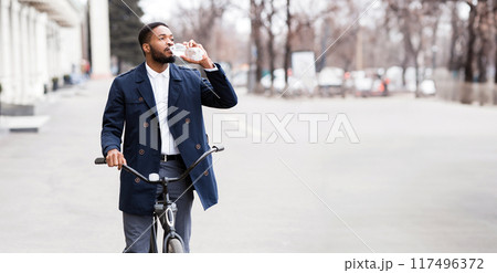 An executive black guy is cycling to the office through downtown, drinking water. The mood is lively and productive, with an urban backdrop highlighting a modern and active commute. An executive black guy is cycling to the office through downtown, drinking water. The mood is lively and productive, with an urban backdrop highlighting a modern and active commute. 117496372