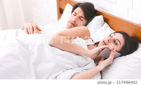 A woman lies in bed, talking on her phone while her partner sleeps soundly beside her. The woman expression suggests she is engaged in a private conversation. 117496402