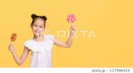 A young girl with pigtails stands holding a colorful lollipop in one hand, while another lollipop is visible in her pocket. She appears joyful and excited about her sweet treats, copy space A young girl with pigtails stands holding a colorful lollipop in one hand, while another lollipop is visible in her pocket. She appears joyful and excited about her sweet treats, copy space 117496429