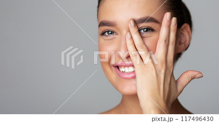 Cheerful young woman covering her face with palm, peeking from fingers and smiling, grey background, panorama with copy space 117496430
