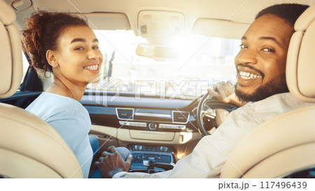 African American young couple sits in their car. The man is driving while the woman is smiling at the camera. They are enjoying their time together and seem happy. 117496439