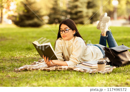 Asian woman is laying on the grass engrossed in a book she is reading. The sun shines overhead as she turns the pages, fully immersed in her literary world. 117496572