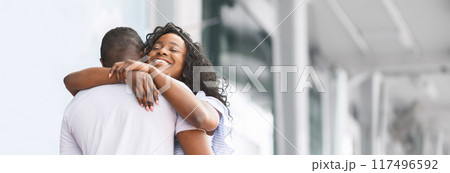 African American woman with long curly black hair is embracing a man. They are standing outside a modern white building with a clear sky in the background, copy space African American woman with long curly black hair is embracing a man. They are standing outside a modern white building with a clear sky in the background, copy space 117496592