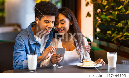 Two African American teenagers are sitting at a cozy cafe, enjoying milkshakes and a slice of cake. They are smiling and watching something on a smartphone together. 117496596