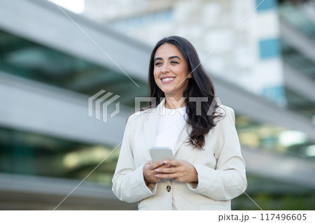 A smiling woman with long dark hair is dressed in a white blazer and a white t-shirt. She is standing in an urban setting with a modern office building in the background, holding her smartphone A smiling woman with long dark hair is dressed in a white blazer and a white t-shirt. She is standing in an urban setting with a modern office building in the background, holding her smartphone 117496605