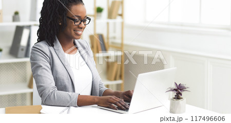 Smiling African American businesswoman using her laptop at her desk in a contemporary office setting, panorama with copy space 117496606