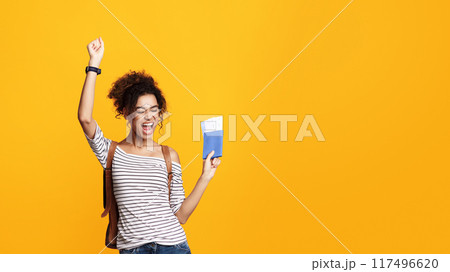 African American young woman with curly hair exudes excitement as she raises one fist in triumph. She is holding a passport and tickets on yellow background, copy space 117496620