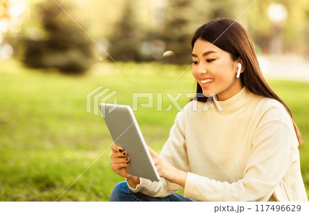 Asian woman is seated on the grass, engaged with a tablet device. She appears focused on the screen, possibly reading or watching something. The grass is green and well-kept 117496629