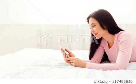 A woman is laying on a bed, focused on a cell phone screen in her hands. She appears engrossed in the device, with a relaxed posture. The room is dimly lit, emphasizing the screens glow. 117496630