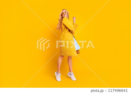 A happy young woman wearing a yellow coat and white sneakers, stands with a bright smile on her face and poses playfully in front of a yellow background. A happy young woman wearing a yellow coat and white sneakers, stands with a bright smile on her face and poses playfully in front of a yellow background. 117496641