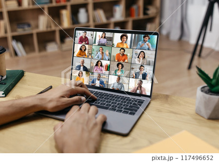 A persons hands are shown typing on a laptop keyboard, the screen of which displays a video conference with multiple participants. 117496652