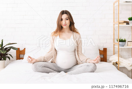 A pregnant woman is sitting cross-legged on a bed, practicing meditation. She has her hands resting on her belly, eyes closed in concentration. 117496716