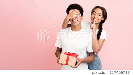 A cheerful African American couple engaged in a playful surprise gift exchange. The girl covers the guy eyes while holding a wrapped present that adds to the excitement of the moment. 117496718