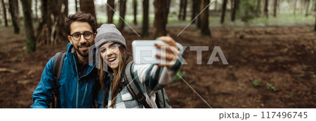 A young couple, dressed in casual outdoor clothing, stands in a forest and takes a selfie with a smartphone. They are smiling and seem to be enjoying their time together, copy space A young couple, dressed in casual outdoor clothing, stands in a forest and takes a selfie with a smartphone. They are smiling and seem to be enjoying their time together, copy space 117496745
