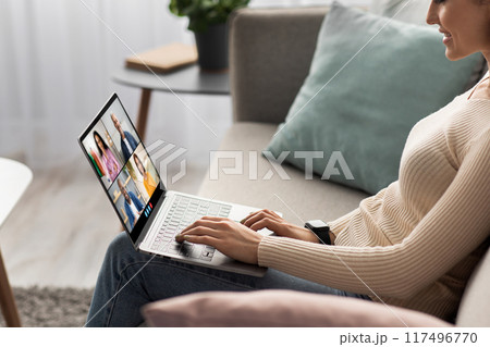 A woman sits on a couch at home and uses a laptop computer to participate in an online video conference meeting. She is smiling and appears engaged in the conversation, side view A woman sits on a couch at home and uses a laptop computer to participate in an online video conference meeting. She is smiling and appears engaged in the conversation, side view 117496770