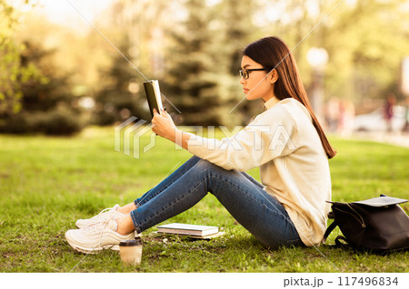 Asian young woman sits on the grass in a serene park engrossed in reading a book. Surrounded by trees, she enjoys the warm, sunny weather with an iced coffee nearby and a backpack by her side. 117496834