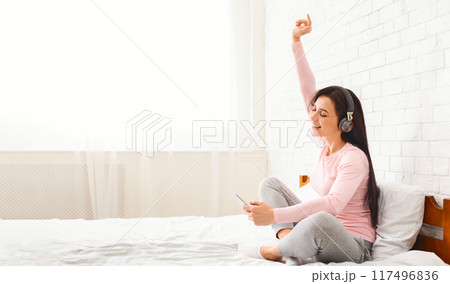 A woman in comfortable loungewear joyfully sings and listens to music on her headphones while sitting on a bed. She holds her smartphone in one hand, raising the other in excitement. 117496836
