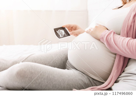 A pregnant woman is resting on a bed, comfortably dressed in casual clothing, while holding an ultrasound picture of her baby. Soft natural light filters through a window in the background, cropped 117496860