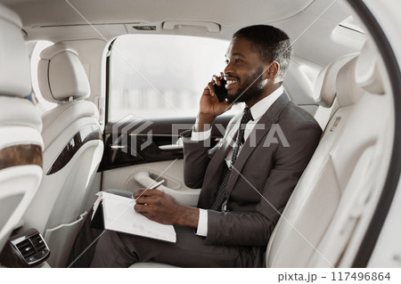Black businessman is sitting in the backseat of a car, taking notes on a notepad while talking on a phone. He is wearing a suit and tie, and he has a serious expression on his face. Black businessman is sitting in the backseat of a car, taking notes on a notepad while talking on a phone. He is wearing a suit and tie, and he has a serious expression on his face. 117496864