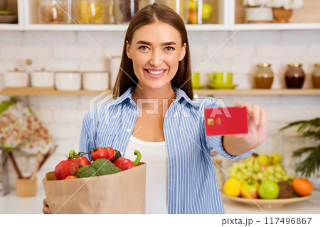 A woman stands in her kitchen, smiling and holding a grocery bag filled with produce and a credit card. A woman stands in her kitchen, smiling and holding a grocery bag filled with produce and a credit card. 117496867