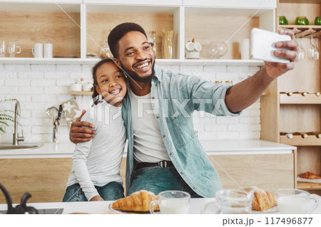 African man and his little girl making selfie with freshly baked croissant, kitchen background, copy space 117496877