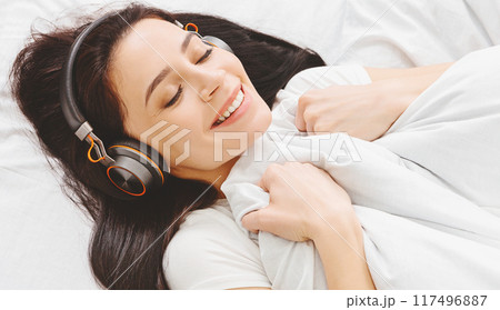 A woman is lying comfortably in bed with headphones on, listening to music or podcasts. She appears relaxed and immersed in the sound coming through the headphones, closeup 117496887