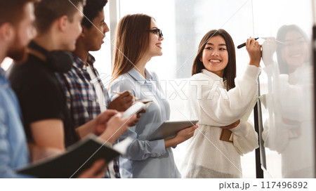 Girl showing answers on whiteboard in classroom with classmates standing around 117496892