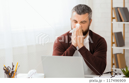 Sick man sitting in front of a laptop is blowing his nose into a tissue. He appears to be dealing with a cold or allergies while using the computer. Sick man sitting in front of a laptop is blowing his nose into a tissue. He appears to be dealing with a cold or allergies while using the computer. 117496909