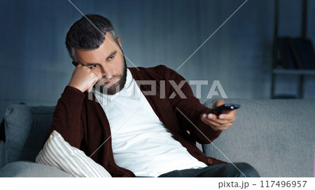 A man with a beard sits on a couch in a dimly lit living room, holding a TV remote with a bored expression on his face. He rests his head on his hand, leaning on a pillow 117496957