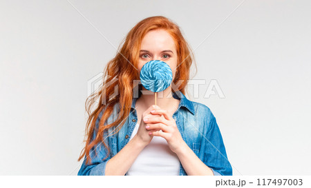 A young woman with red hair stands against a light gray background, holding a blue lollipop in front of her mouth as if it were a makeshift microphone 117497003